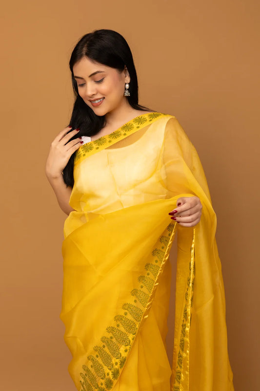 Woman wearing a yellow saree with green border against a brown background